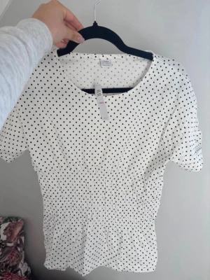 Vit prickig topp med smock  - Söt vit topp från Lindexmed svarta prickar och rund halsringning. Toppen har korta ärmar och smockad midja som ger en snygg passform. Perfekt till jeans eller kjol för en trendig look. Ny med prislapp, storlek 158/164. Passar xs-m på grund av stretchen