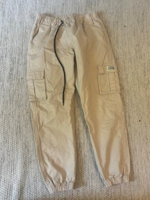 Beiga cargobyxor med resår och fickor - Säljer ett par beiga cargobyxor från Jack & Jones med resår i midjan och benslut, samt snörning i midjan. Byxorna har stora sidofickor och bakfickor, perfekta för en avslappnad och trendig look. 
