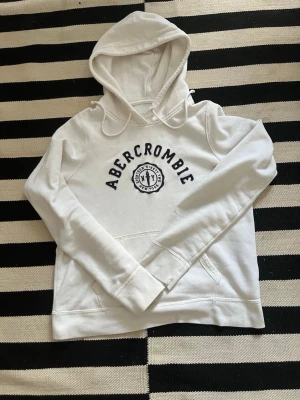 Vit hoodie från Abercrombie - Snygg vit hoodie från Abercrombie med svart tryck på bröstet och klassisk känguruficka. Hoodien har dragsko i huvan och ribbade muddar vid ärmslut och nederkant. Perfekt för en avslappnad och trendig look. Storlek M men passar S och XS.