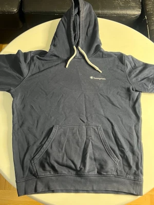 Blå hoodie från Champion - Mörkblå hoodie från Champion med vit broderad logga på bröstet och vita dragsnören i huvan. Klassisk känguruficka framtill och ribbade muddar. Perfekt för en avslappnad och sportig stil.