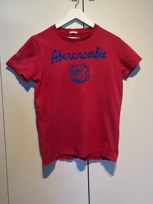 Röd Abercrombie t-shirt med brodyr - Snygg röd t-shirt från Abercrombie med blå broderad text och bulldog på bröstet. Klassisk rund hals och korta ärmar. Tillverkad i mjuk bomull som känns skön mot huden. Perfekt för en avslappnad och cool stil.