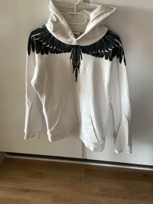 Vit Marcelo Burlon hoodie - Säljer en vit hoodie från Marcelo Burlon med ikoniskt svart vingmönster över axlar och bröst. Hoodien har huva, känguruficka och är tillverkad i mjuk bomull. Perfekt för dig som vill sticka ut med en unik och trendig look.
