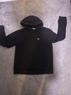 Svart hoodie från Lacoste - Svart hoodie från Lacoste med klassisk krokodillogga på bröstet. Tröjan har huva och en stor magficka framtill.  ❌ Inga defekter på tröjan.