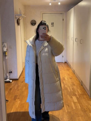 Lång vit dunjacka med bälte - Säljer en lång, vit dunjacka med bälte i midjan och hög krage. Jackan har quiltad design och dragkedja framtill. Perfekt för kalla vinterdagar och riktigt snygg att styla med jeans eller boots.