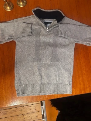 Tommy Hilfiger half zip - Snygg grå Tommy hilfiger tröja med half zip och hög krage. Stor H-detalj framtill och ribbade muddar vid ärmslut och nederkant. Perfekt för dig som gillar klassisk och enkel stil. Priset kan diskuteras 