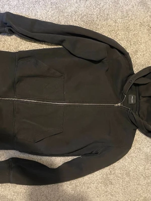 Svart zip hoodie med huva - En klassisk svart hoodie från Jack & Jones med dragkedja framtill och två stora fickor. Hoodien har en mjuk insida och en rymlig huva. Perfekt för chill dagar eller när du vill ha en enkel och clean look.