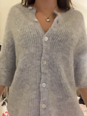 Ljusgrå stickad kofta med knappar - Mysig ljusgrå stickad kofta med ribbad struktur och vita knappar framtill. Koftan har halvlånga ärmar och rund halsringning. Materialet ser fluffigt och mjukt ut, perfekt för lager på lager och chill dagar.