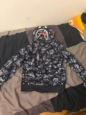 BAPE Shark camo hoodie svart/grå - Säljer en BAPE Shark hoodie med ikoniskt hajmotiv på huvan och svart/grått camomönster. Dragkedja framtill och ribbade muddar. Materialet är mjukt och bekvämt, perfekt för streetwear-stilen. Insidan är ljus och slät. Grym hoodie för dig som vill sticka ut!