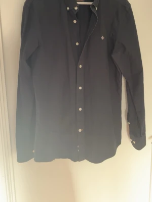 Mörkblå skjorta med vita knappar - Stilren mörkblå skjorta med klassisk krage och vita knappar. Skjortan har lång ärm och en liten broderad logga på bröstet. Perfekt till både jeans och chinos för en clean look.