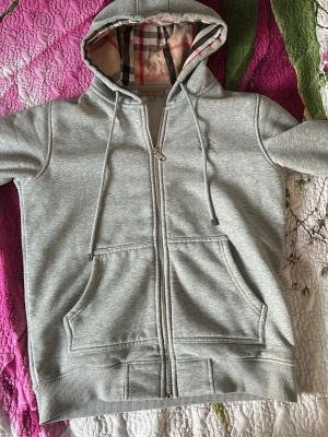 Grå zip hoodie från Burberry - Snygg grå zip hoodie från Burberry med klassiskt rutigt foder i huvan. Hoodien har dragsko i huvan, två stora fickor fram och ribbade muddar. Tillverkad i mjukt bomullsmaterial som känns skönt mot huden. Perfekt för en avslappnad och stilren look.