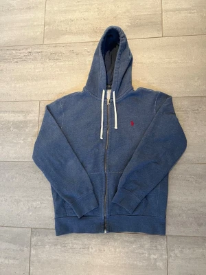 Blå zip hoodie från Polo Ralph Lauren - Blå hoodie med dragkedja från Polo Ralph Lauren. Klassisk design med vit dragsko i huvan och röd broderad logga på bröstet. Tillverkad i mjukt bomullsmaterial och har två fickor framtill. Perfekt för en avslappnad och stilren look.
