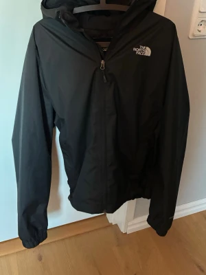 Svart vindjacka från The North Face - Snygg svart vindjacka från The North Face med huva och dragkedja framtill. Jackan har logga på bröstet och är tillverkad i ett lätt, vindtätt material. Perfekt för blåsiga dagar och enkel att matcha med olika outfits.