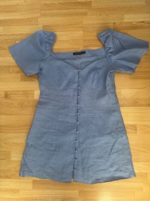 Ny. Ljusblå kortklänning med puffärm - Supersöt ljusblå kortklänning från Zara med puffiga kortärmar och knappar framtill. Klänningen har en feminin passform och är tillverkad i ett svalt linnematerial, perfekt för varma dagar. Fyrkantig ringning ger en trendig touch.Linne 100%. Klänningen köpte jag på rea pga blekt färg, se sista bilden.