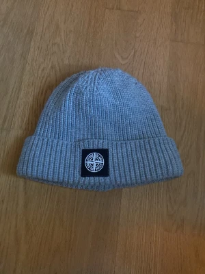 Grå ribbstickad mössa Stone Island - Säljer en grå ribbstickad mössa från Stone Island med uppvikt kant och klassisk svart patch med logga framtill. Mössan är i mjukt material och har en enkel, clean look som passar till det mesta.