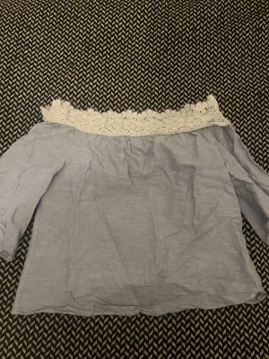 Blå offshoulder topp med spetskant - Söt blå offshoulder topp med vit spetskant upptill. Toppen har trekvartsärm och är tillverkad i ett lätt bomullstyg med diskret randigt mönster. Perfekt för dig som gillar romantiska detaljer och en avslappnad stil.