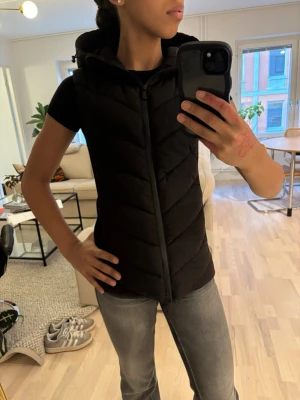 Svart dunväst med huva (Zara väst) - Snygg svart dunväst med huva och dragkedja framtill. Västen har ett quiltat mönster i diagonala ränder och är lätt vadderad för extra värme. Perfekt att slänga över en t-shirt eller hoodie för en casual look. Använd få gånger💕