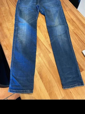 Blå raka jeans med slitningar - Säljer ett par klassiska blå jeans med raka ben och snygga slitningar framtill. Replay Anbass Hyperflex Jeansen har fem fickor och kontrastsömmar. Perfekta för en avslappnad och trendig look. Materialet är denim i bomull. I mycket bra skick, sparsamt använda. 30:30 i storlek 