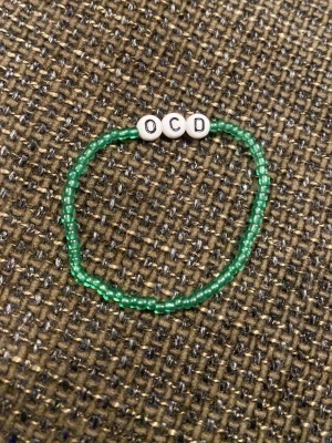 Grönt pärlarmband med bokstäver - Ett handgjort armband med små gröna pärlor och tre vita pärlor med svarta bokstäver som stavar 'OCD'. Armbandet har en enkel och ungdomlig design, perfekt för dig som gillar personliga accessoarer.