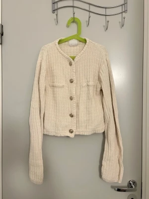 Beige stickad kofta med knappar - Säljer en beige stickad kofta med rund halsringning och dekorativa fickor framtill. Koftan har långa ärmar och stängs med fem stora knappar. Perfekt att slänga över en t-shirt för en chill look.