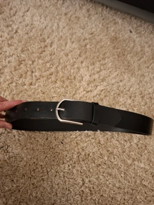 Svart klassiskt bälte i skinn - Snyggt svart bälte i skinn med enkel, silverfärgad metallspänne. Bältet har flera hål för justerbar passform. Säljer pga för liten storlek.