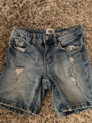 Ljusblå denimshorts från 157 - Säljer ett par ljusblå denimshorts från 157 med slitna detaljer och femficksmodell. Shortsen har normal passform, bälteshällor och klassisk knappgylf. Perfekta för sommaren och ger en avslappnad look.