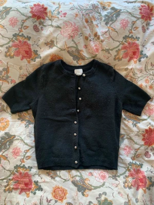 Svart kortärmad kofta med pärlknappar - Snygg svart kortärmad kofta med pärlknappar framtill. Säljer då den knappast kommit till användning, väldigt bra skick