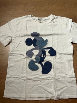Vit Mickey Mouse t-shirt Stradivarius  - Vit t-shirt från Stradivarius med stor blå Mickey Mouse-figur på framsidan och texten 'ALWAYS ORIGINAL Mickey Mouse' i marinblått på ryggen. Klassisk passform och korta ärmar, perfekt för dig som gillar ikoniska tryck och streetstyle.