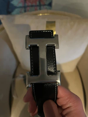 Svart skärp med silverfärgat H spänne - Snyggt svart bälte med ett stort, silverfärgat H format spänne. Bältet har vita sömmar längs kanterna och är gjort i slätt skinn. Perfekt för att lyfta vilken outfit som helst.