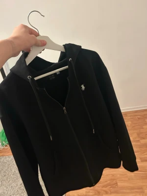 Svart zip hoodie från Polo Ralph Lauren - Säljer en svart hoodie med dragkedja från Polo Ralph Lauren. Klassisk design med huva, dragsnören och fickor framtill. Ikoniska vita loggan broderad på bröstet. Tillverkad i mjukt bomullsmaterial som är skönt att ha på sig.