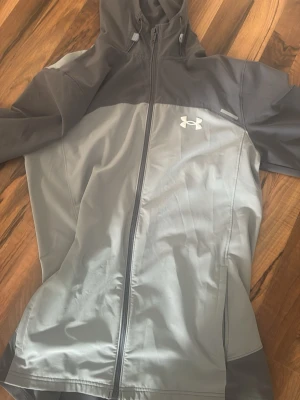 Grå vindjacka från Under Armour - Snygg grå vindjacka från Under Armour med huva och dragkedja framtill. Jackan har två olika grå nyanser, logga på bröstet och stort Under Armour-tryck på ryggen. Perfekt för dig som vill ha en sportig och stilren look.jätte bra skick 