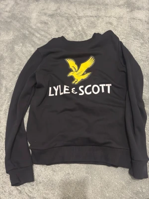 Lyle & Scott tröja - Svart stickad tröja från Lyle & Scott med stor gul logga och text på ryggen. Tröjan har ribbade muddar vid ärmslut och nederkant, samt en klassisk rund halsringning. Perfekt för dig som gillar streetwear och tydliga loggor.