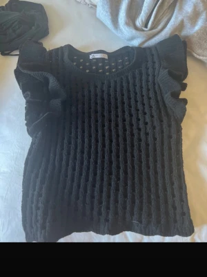 Svart stickad topp med volangärmar - Snygg svart stickad topp från Bershka med coolt hålmönster och volangdetaljer på axlarna. Rund halsringning och korta ärmar ger en trendig look. Perfekt att styla lager på lager eller som statement-piece.