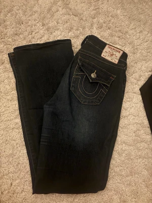 True Religion Becca Bootcut Jeans - Säljer ett par mörkblå bootcut jeans från True Religion, modell Becca Mid Rise Bootcut. Jeansen har snygga kontrastsömmar, klassisk fickdesign med lock och knapp bak samt en ikonisk logga på bakfickan. Tillverkade i slitstark denim med skön passform.