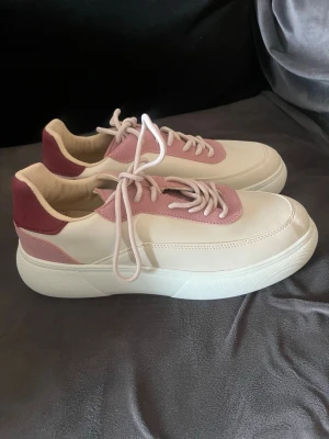 Krämvita och rosa sneakers - Snygga sneakers i krämvitt med rosa detaljer och vinröd häl. Skorna har tjock vit sula, rosa snörning och ovandel i syntetmaterial med mockaliknande parti bak. Perfekta för dig som gillar en stilren men ändå färgglad look. Nya så i perfekt skick