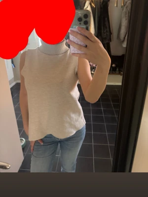 Beige ärmlös polotopp - Säljer en stilren beige polotopp utan ärmar. Toppen har en hög ribbad krage och är gjord i ett mjukt, stickat material. Perfekt att matcha med jeans eller kjol för en clean look.