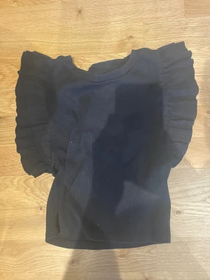 Svart topp med volangärmar💕 - Trendig svart topp med breda volangärmar och rund halsringning. Toppen har en croppad passform och är tillverkad i ett ribbat material som ger en snygg struktur. Perfekt för dig som vill ha en statement-topp till din outfit.💕