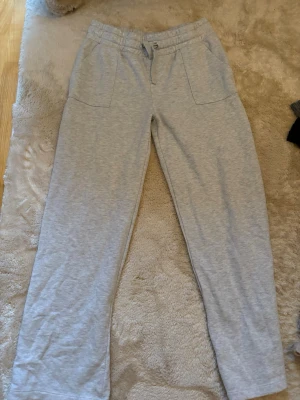 Ljusgrå mjukisbyxor med raka ben - Säljer ett par ljusgrå mjukisbyxor med raka ben och resår i midjan. Byxorna har två stora fickor fram och två bakfickor. Perfekta för chill dagar hemma eller när du vill vara bekväm. Materialet känns mjukt och skönt mot huden.