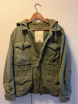 Olivgrön vindjacka med huva - Snygg olivgrön vindjacka från Being Supreme med fyra stora fickor framtill, dragsko i midjan och huva med snören. Jackan har knapp- och dragkedjestängning samt justerbara ärmslut. Perfekt för dig som gillar streetwear och funktionell stil.