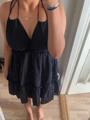 Svart kort volangklänning med smala band - Svart kort klänning med smala axelband med v-ringning. Klänningen har volangdetaljer och en ”luftig” passform. Materialet känns lätt och bekvämt mot huden. Använt 2 gånger