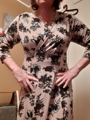 Cappuccino färgad blommig omlottklänning från Cotton Traders  - Snygg cappuccino färgad långklänning med svart blommigt mönster och omlottdesign. Klänningen har v-ringning och trekvartsärm, vilket ger en avslappnad men ändå chic look. Perfekt för dig som gillar feminina detaljer och mjuka material. Storlek UK 16, EU 44/46/XL. Mått på bilderna. 