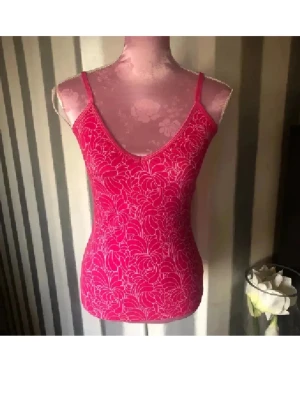 Rosa blommig linnetopp med smala axelband - Säljer en snygg rosa linnetopp med blommigt mönster och smala axelband. Toppen har en v-ringad front och är figurnära med en ljusrosa blommig design över hela tyget. Perfekt för dig som gillar färg och mönster. Med inbyggd bh 
