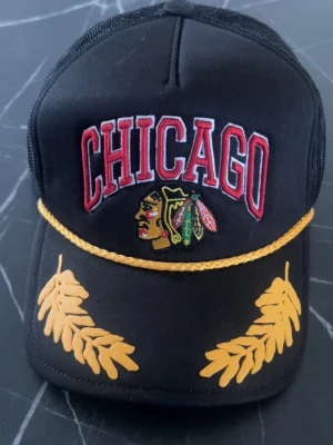 Svart Chicago keps med brodyr - Snygg svart keps med broderad CHICAGO-text i rött och vitt, samt klassisk Blackhawks-logga framtill. Guldigt snöre och broderade blad på skärmen ger extra detaljer. Justerbar baksida och mesh-paneler för skön passform.