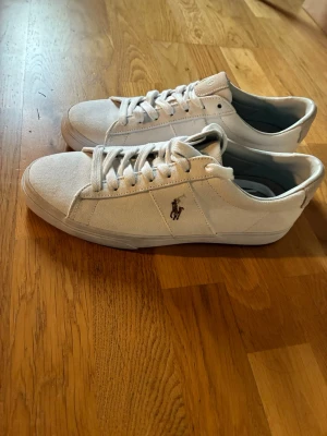Vita Polo Ralph Lauren canvas sneakers - Snygga vita sneakers från Polo Ralph Lauren i canvasmaterial med klassisk låg profil och den ikoniska broderade loggan på sidan. Skorna har vita snören och en stilren design som passar till det mesta. Perfekta för dig som gillar enkel och clean stil.