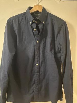 Mörkblå skjorta från Selected Homme - Klassisk mörkblå skjorta från Selected Homme med button-down krage och bröstficka. Skjortan har långa ärmar och vita knappar framtill. Perfekt till jeans eller chinos för en clean och stilren look.