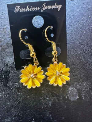Gula blomörhängen i metall - Snygga örhängen i form av gula blommor med guldfärgade detaljer. Blommorna har en glittrande sten i mitten och hänger på krok. Perfekta för att ge din look en färgklick och en lekfull touch.