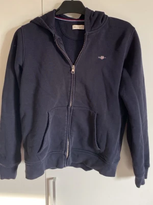 Mörkblå zip hoodie från Gant - Mörkblå hoodie från Gant med dragkedja framtill och fickor på magen. Klassisk huva och liten broderad logga på bröstet. Tillverkad i mjukt sweatshirtmaterial, perfekt för en avslappnad stil. Passar även en person som är 170 storlek 158-164
