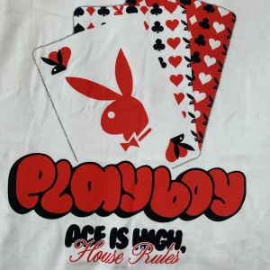 Vit Playboy t-shirt med spelkort - Vit t-shirt från Playboy med stort tryck av spelkort i rött, svart och vitt samt den klassiska Playboy-kaninen. Texten 'Playboy Ace is High, House Rules' i grafisk stil under motivet. Perfekt för dig som gillar statement-plagg och coola prints.