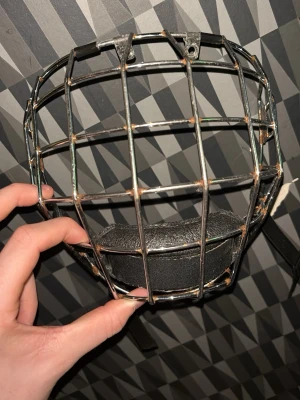 Ishockeygaller i metall med hakskydd - Säljer ett klassiskt ishockeygaller i silverfärgad metall med svart hakskydd i vadderat konstläder. Gallret har fyrkantigt rutmönster och fästen upptill för montering på hjälm. Perfekt för dig som behöver extra skydd på isen. Lite rostigt på vissa ställen