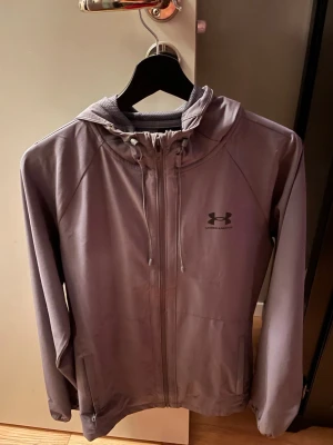 Grå vindjacka från Under Armour - Snygg grå vindjacka från Under Armour med huva och dragkedja framtill. Jackan har snörning i huvan, två sidofickor och är tillverkad i ett lätt, vattenavvisande material. Perfekt för blåsiga dagar och sportiga outfits. Köpt för några månader sen, använd 2gånger. Nypris 1000kr