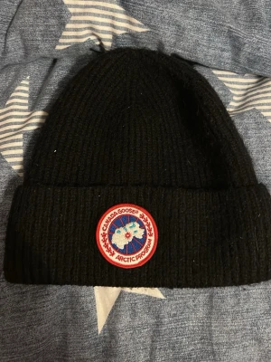 Svart ribbstickad mössa Canada Goose - Säljer en svart ribbstickad mössa från Canada Goose med klassisk rund logotyp framtill i rött, vitt och blått. Mössan har uppvikt kant och är tillverkad i ett tjockt, värmande material som passar perfekt för kalla dagar.